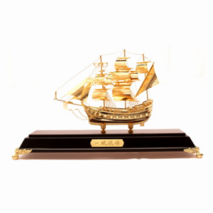 A golden wealth ship model intricately crafted with detailed sails, masts, and decorative elements, displayed on a dark wooden base with gold accents. The ship symbolizes prosperity, financial success, and the attraction of good fortune. It is a popular Feng Shui item used to enhance wealth and bring auspicious energy. The base features an engraved gold plaque, adding a touch of luxury and elegance to this powerful symbol of abundance.