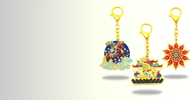 The image shows three keychains with distinct designs, each attached to a gold-colored chain and keyring. The first keychain features a blue and gold design, the second shows a figure resembling a mythical or spiritual creature, and the third has a bright sun-like pattern. The keychains are arranged against a light gradient background.