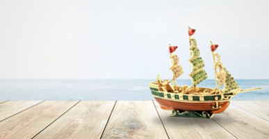 A small, intricately bejeweled ship model with golden details and decorative sails, placed on a wooden surface against a serene seascape background.