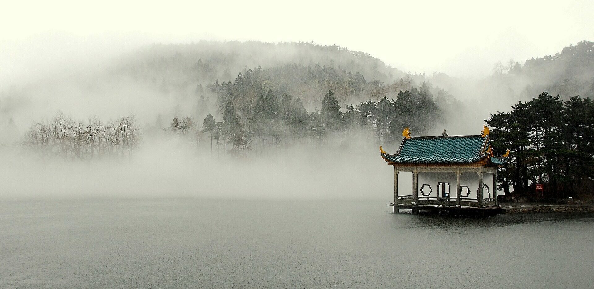 A traditional Chinese pavilion with a green roof and gold accents stands on the edge of a calm lake surrounded by mist. The background features fog-covered trees and rolling hills, creating a serene and mysterious atmosphere.