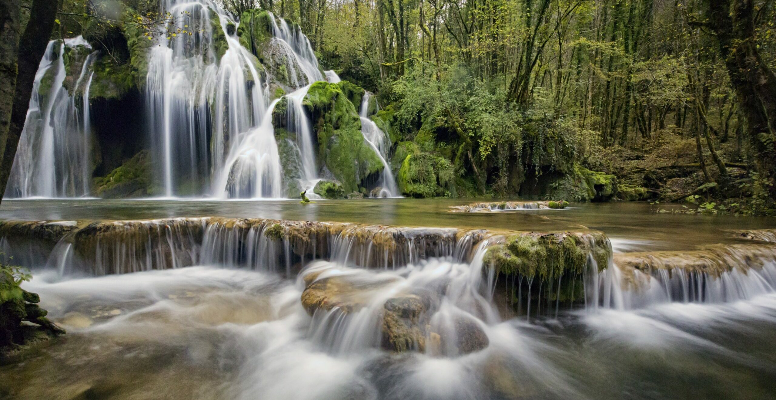 A serene waterfall cascading over moss-covered rocks in a lush green forest. The water flows gently, creating soft, silky textures as it moves through various tiers, surrounded by dense trees and foliage.