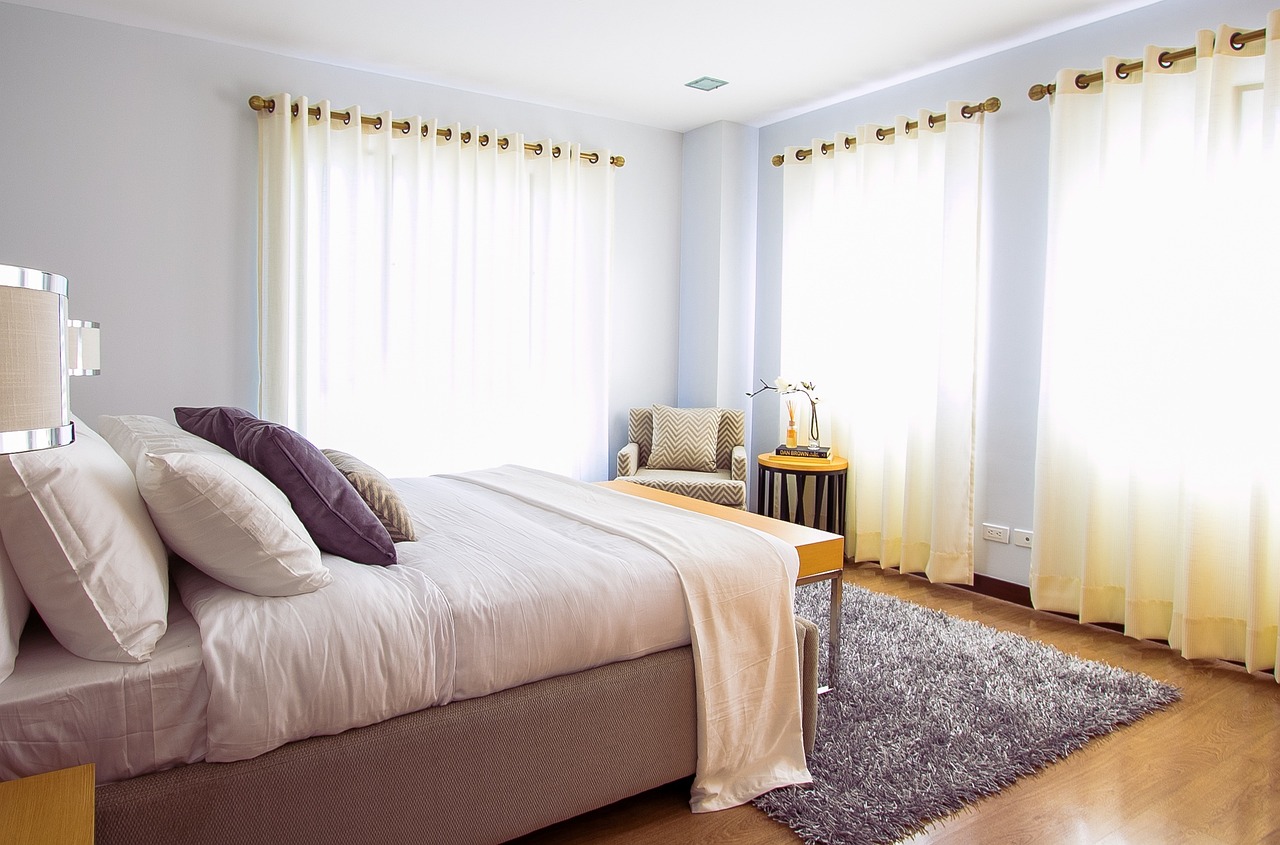 A bright and airy bedroom with large windows covered by sheer white curtains, a neatly made bed with multiple pillows, a light-colored blanket, and a plush rug on the wooden floor. A cozy chair and a small round table with decorative items sit in the corner of the room, enhancing the serene and calm atmosphere.