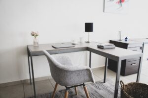 Minimalist L-shaped office desk setup with a gray fabric chair, a small table lamp, a vase with flowers, a laptop, a cup, and a notebook. The workspace is clean and modern with neutral tones, creating a calm and organized environment.