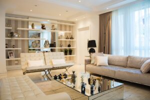 A modern living room with an elegant and sophisticated design. The room features a neutral color palette with beige, cream, and light brown tones. A large beige sofa with white and pastel cushions is situated on the right side, while two cream-colored leather chairs with metal frames are positioned in the center. A glass coffee table with mirrored sides and decorative items, such as small sculptures and glass objects, is placed in front of the seating area.